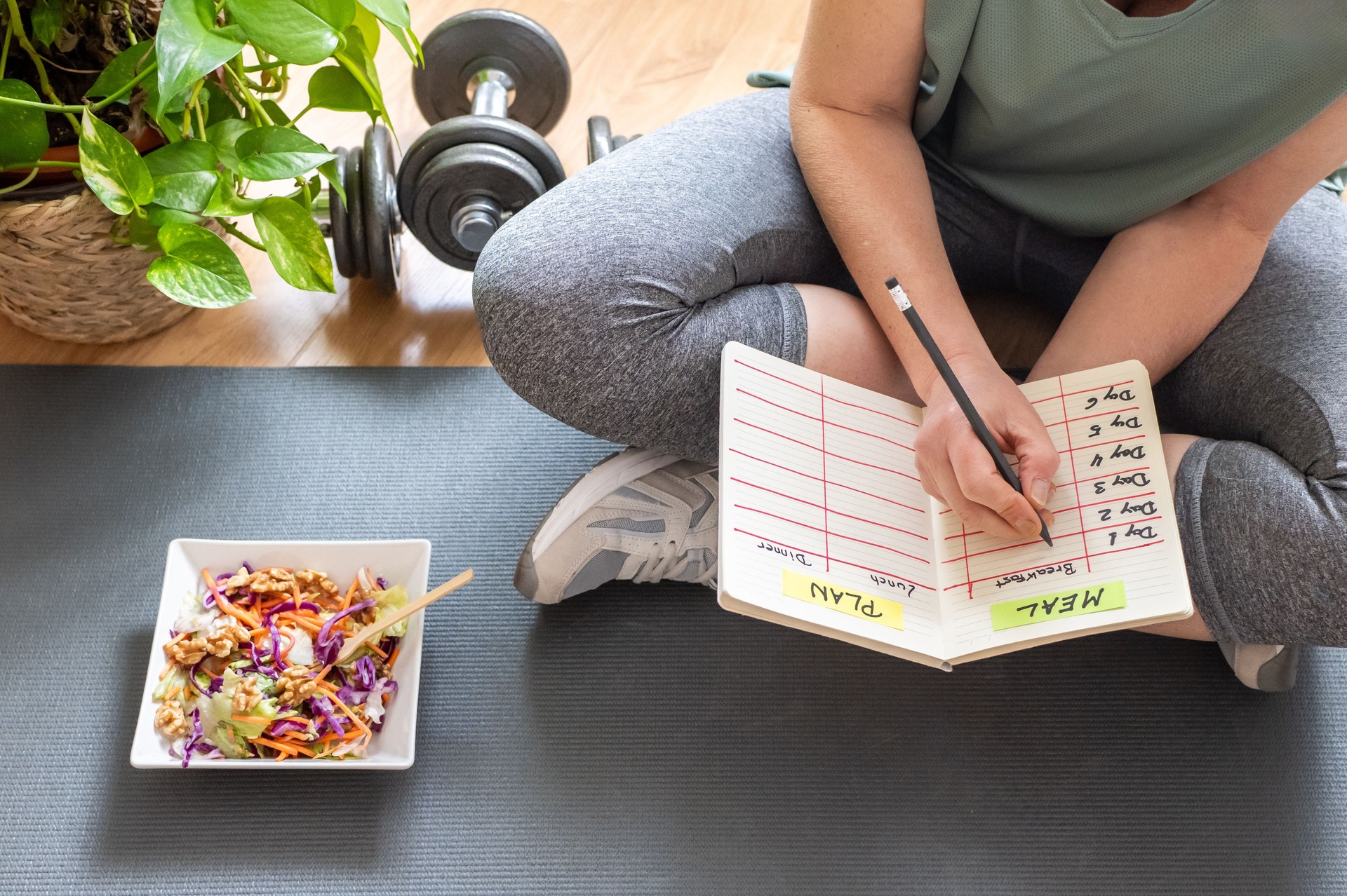 Mature woman sitting on a yoga mat, jotting down her meal plan after completing a workout at home. Nearby, dumbbells and a vibrant salad emphasize her commitment to a healthy lifestyle